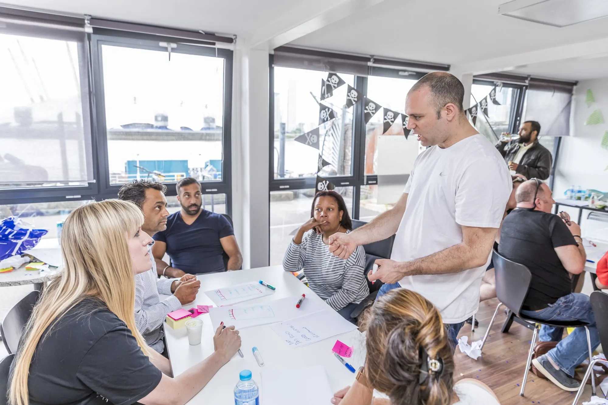 A man in a white T-shirt talks with a table of people by the windows