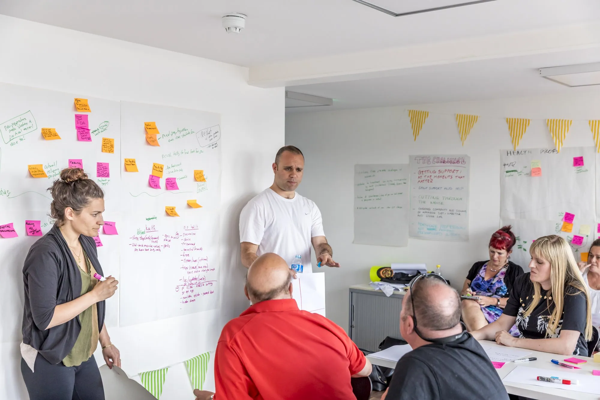 A woman by a wall of sticky notes talks to the group, a man in a white T-shirt beside her