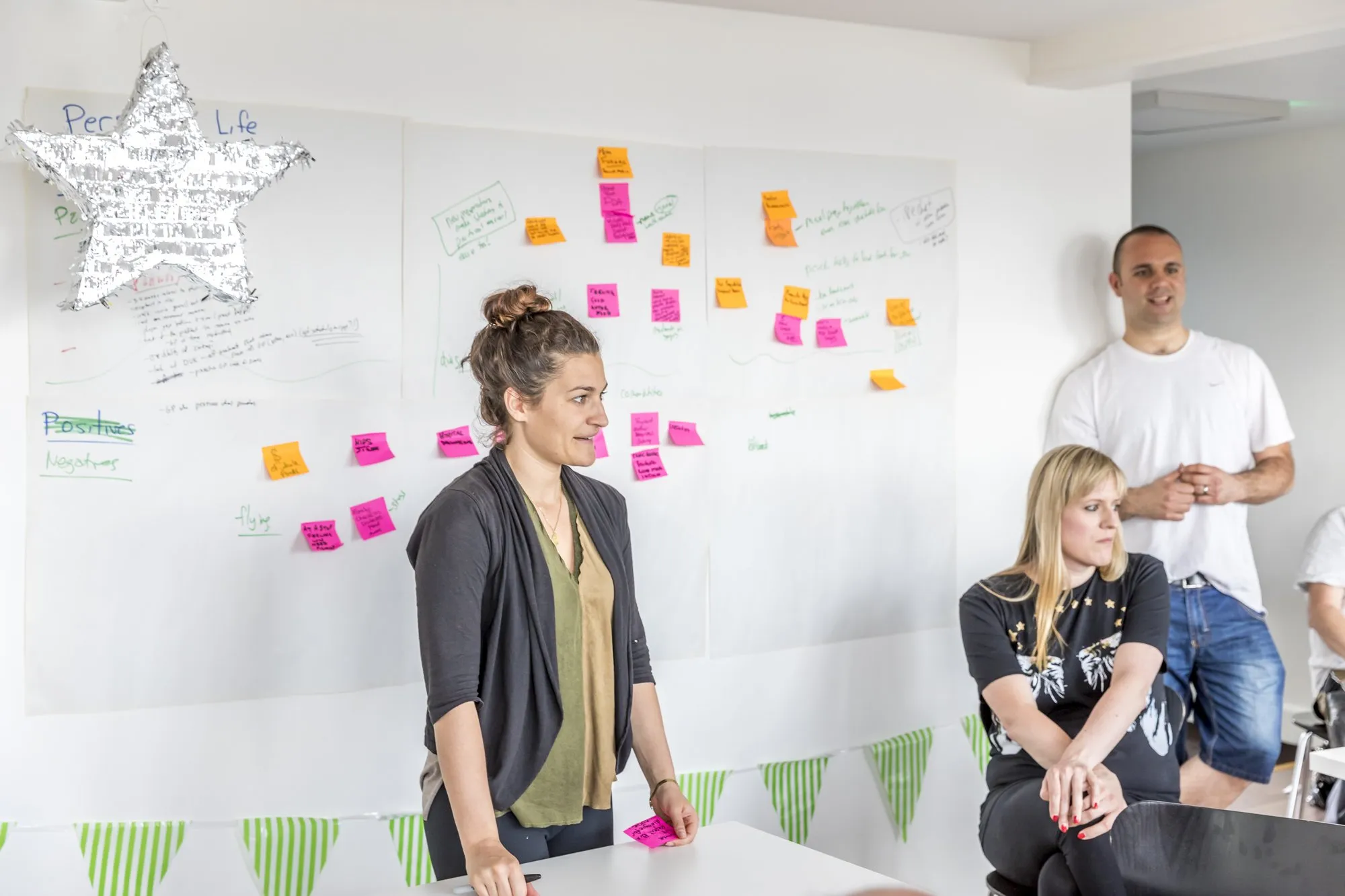 A woman in a dark cardigan talks in front of a wall of sticky notes as two others listen
