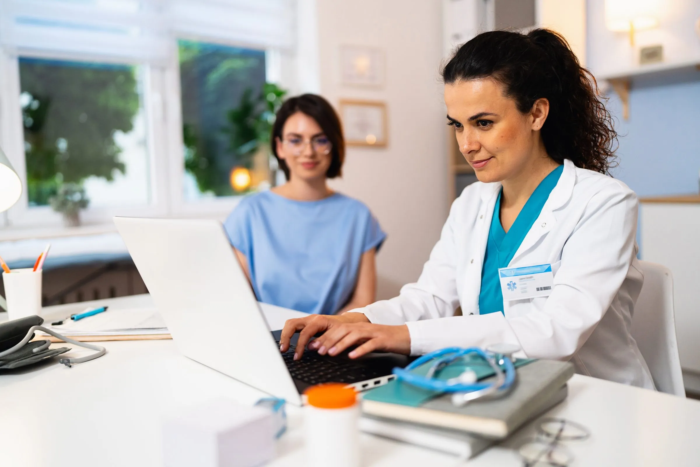A doctor types notes on a laptop as she talks with a patient