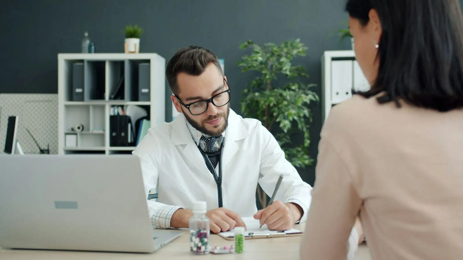 A doctor takes notes at his laptop while talking with a patient