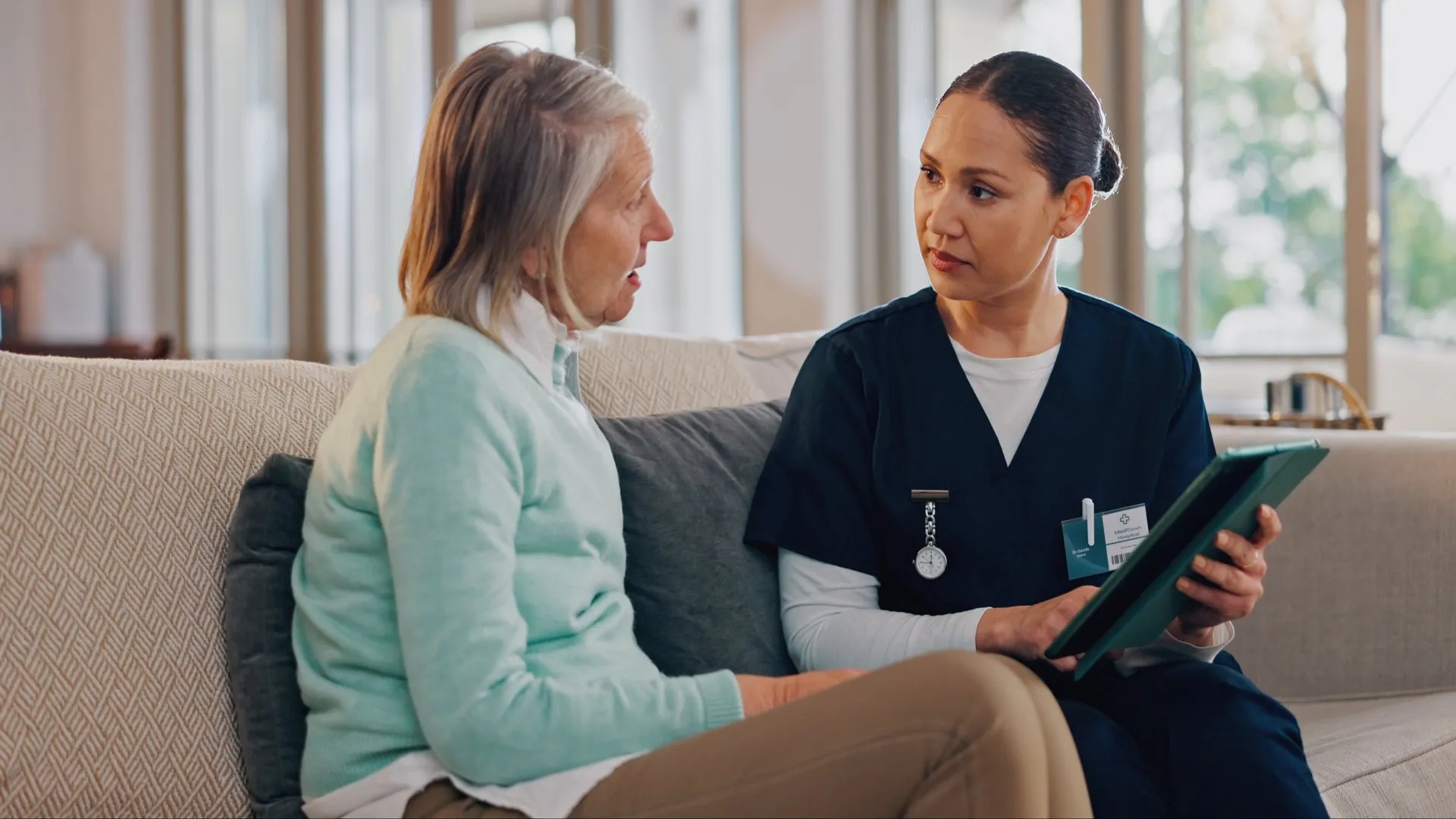 A nurse talks with an older woman at home, a tablet in her hand