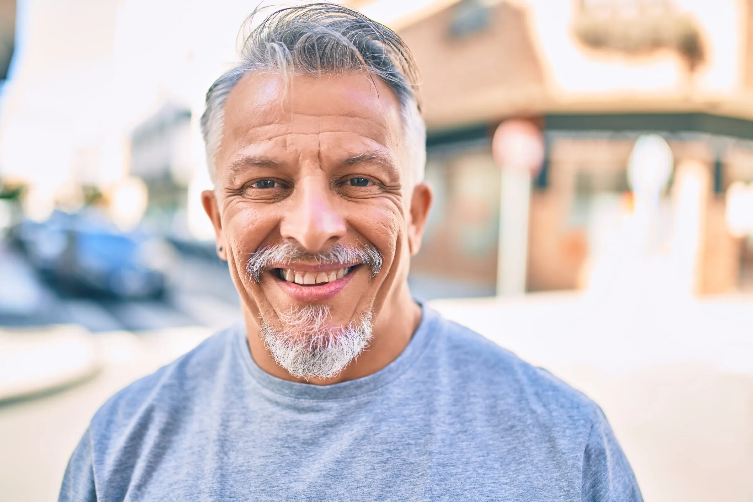 A grey-haired man with a beard smiles on a city street