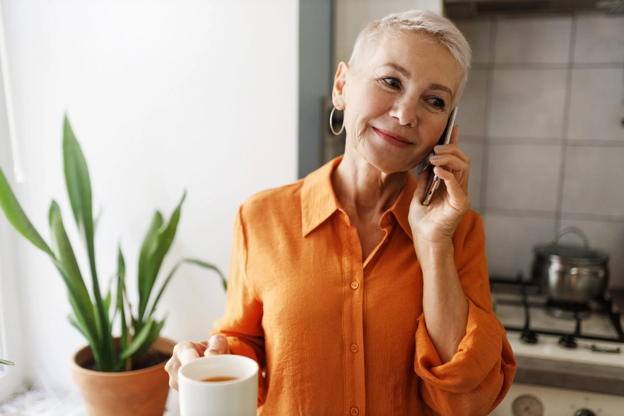 An older woman talks on the phone in her kitchen, holding a cup of tea