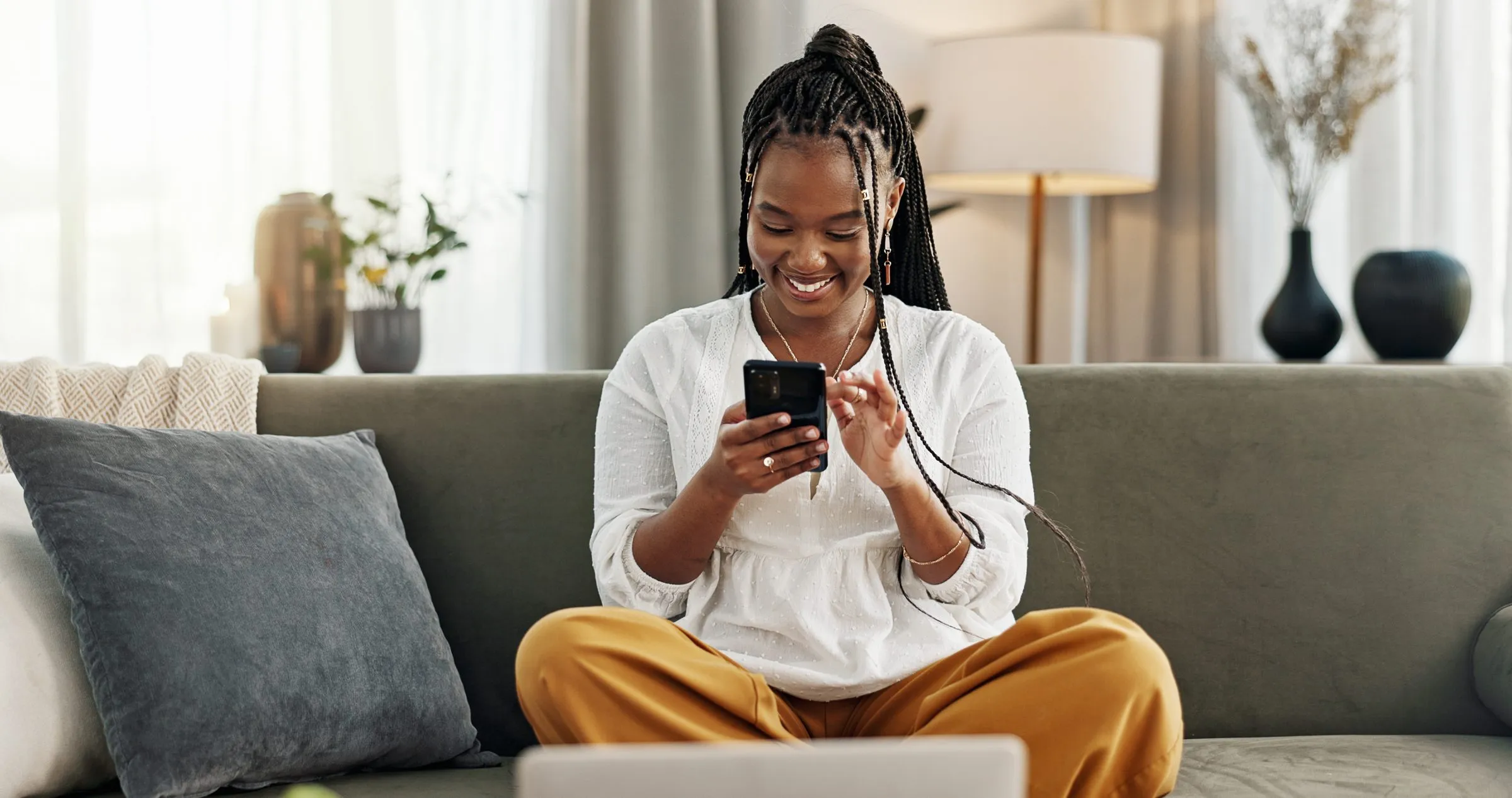 A woman laughs at her phone, sitting cross-legged on her sofa
