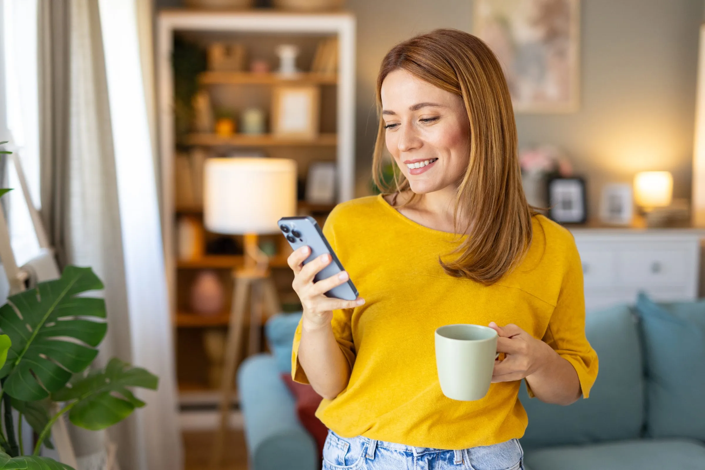 A woman smiles at her phone, holding a mug of tea at home