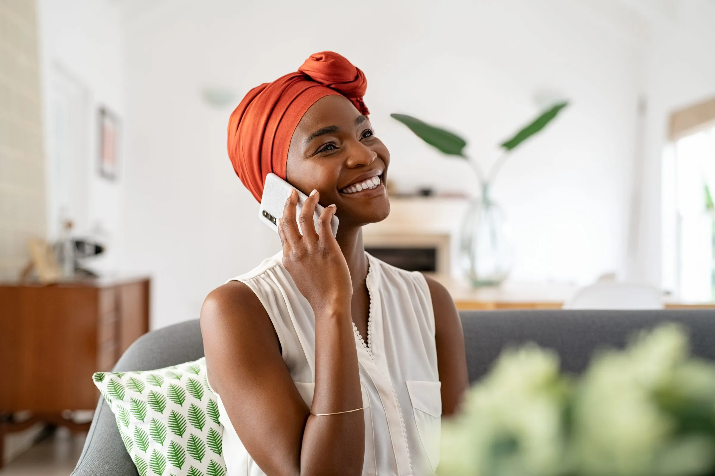 A woman in an orange headwrap smiles as she talks on the phone at home